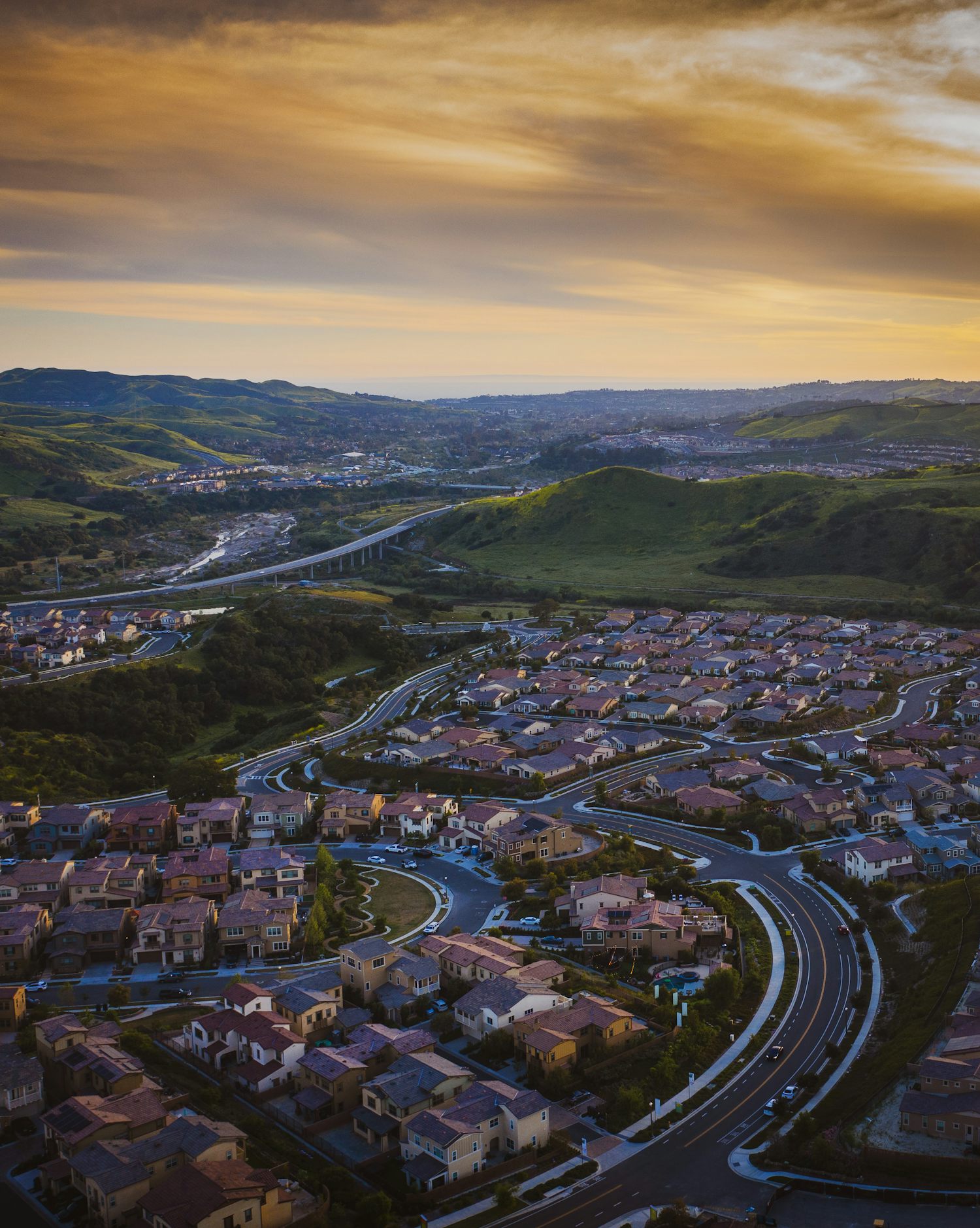 Aerial view of a master-planned residential community at golden hour, rooftops winding along green hillsides toward the coast.